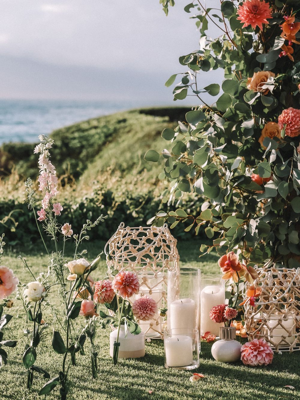 Garden wedding decor with coral dahlias, white lanterns, and candles on grassy hillside.