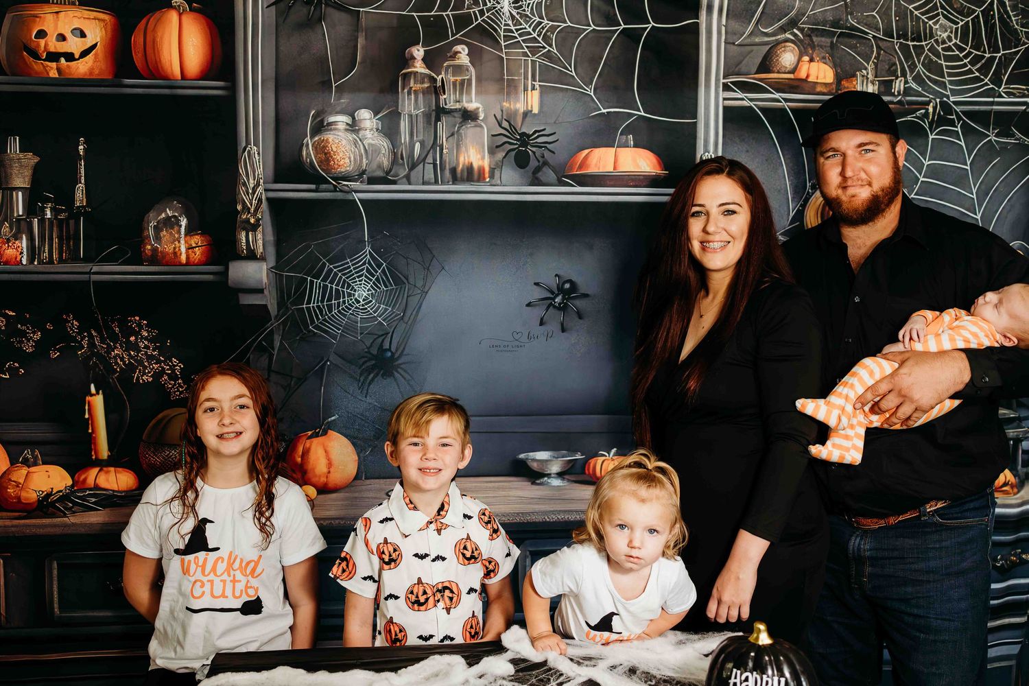 A family poses in front of a Halloween decorated backdrop with pumpkins and spiderwebs.