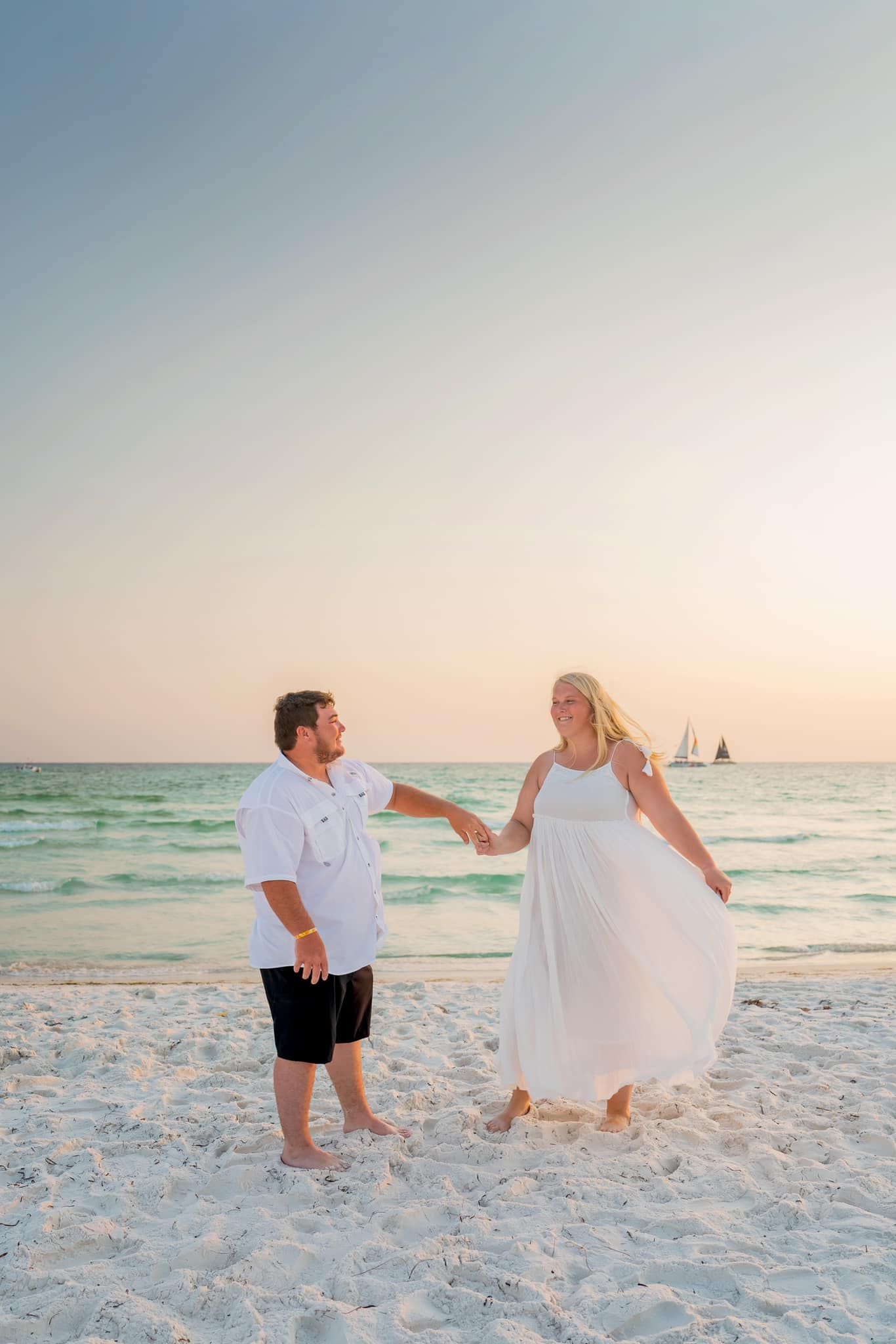 Romantic couple holding hands on a white sand beach at sunset with sailboats in the background.
