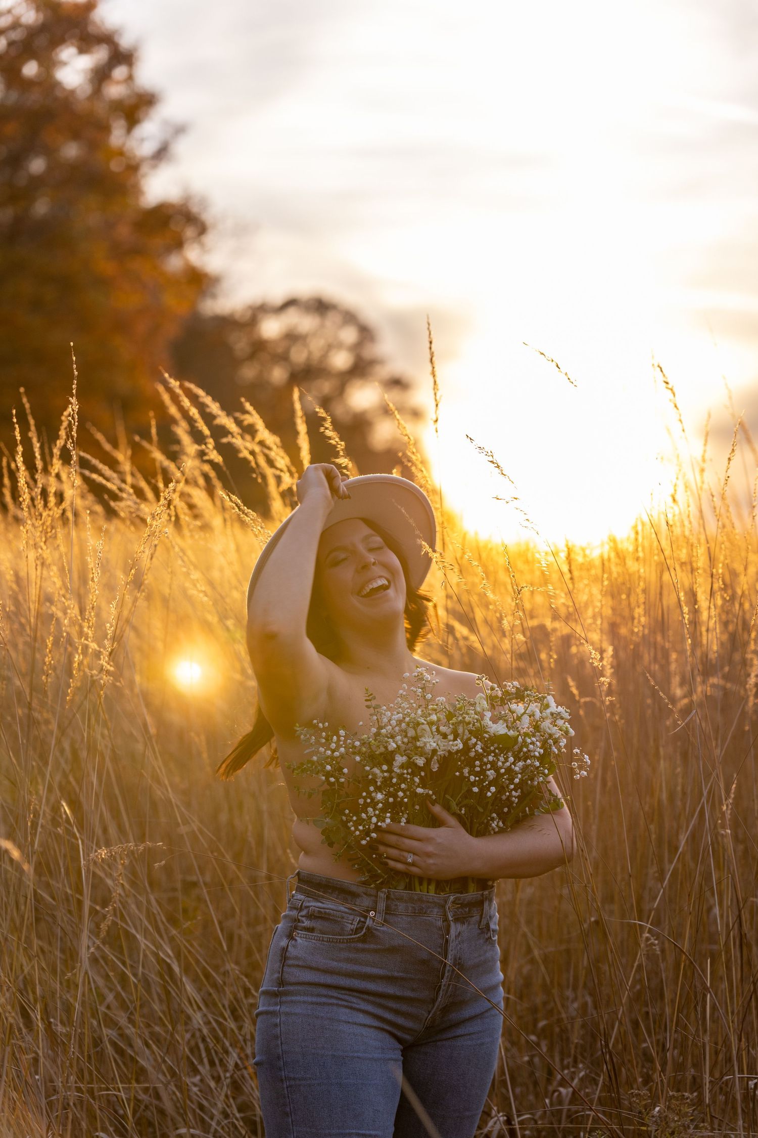Joyful woman laughing in golden field at sunset holding white wildflowers and wearing a wide-brim hat.