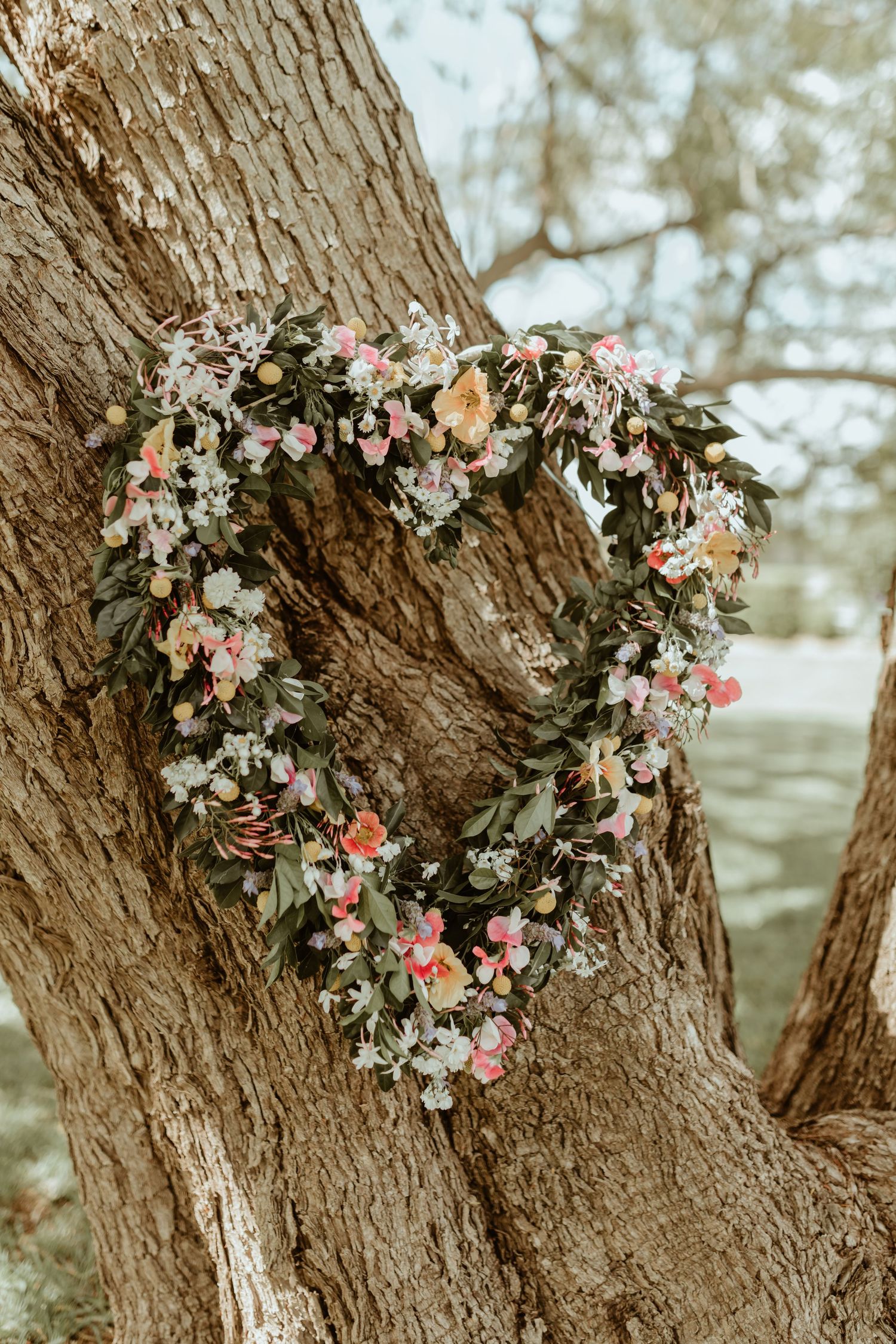 A heart-shaped floral wreath with pink and white flowers hangs on a rustic tree trunk.