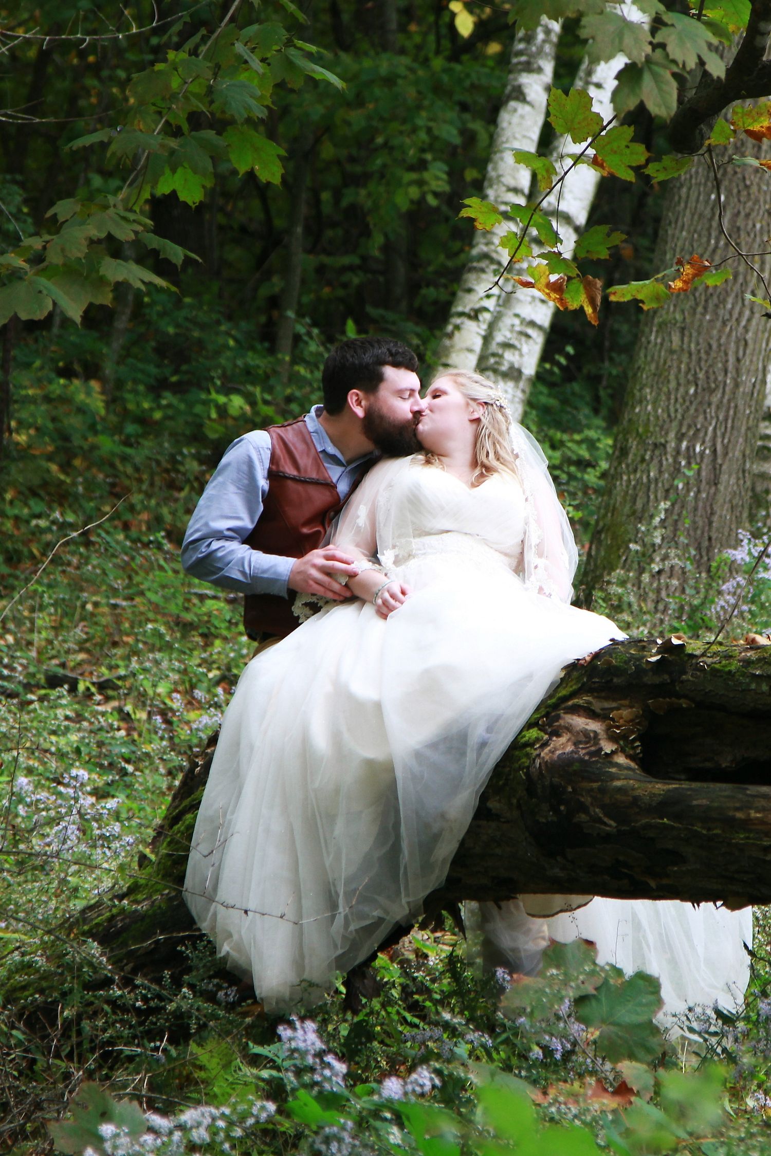 Bride and groom sharing a kiss on a fallen log in lush forest.