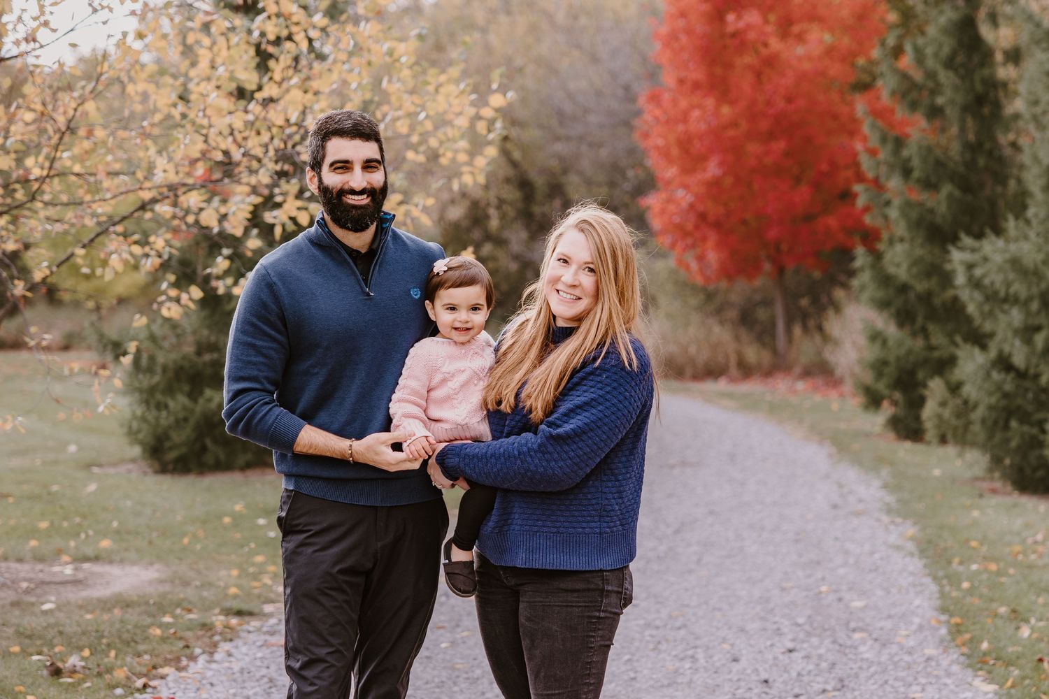 A family stands together on a gravel path surrounded by autumn trees with vibrant red leaves.