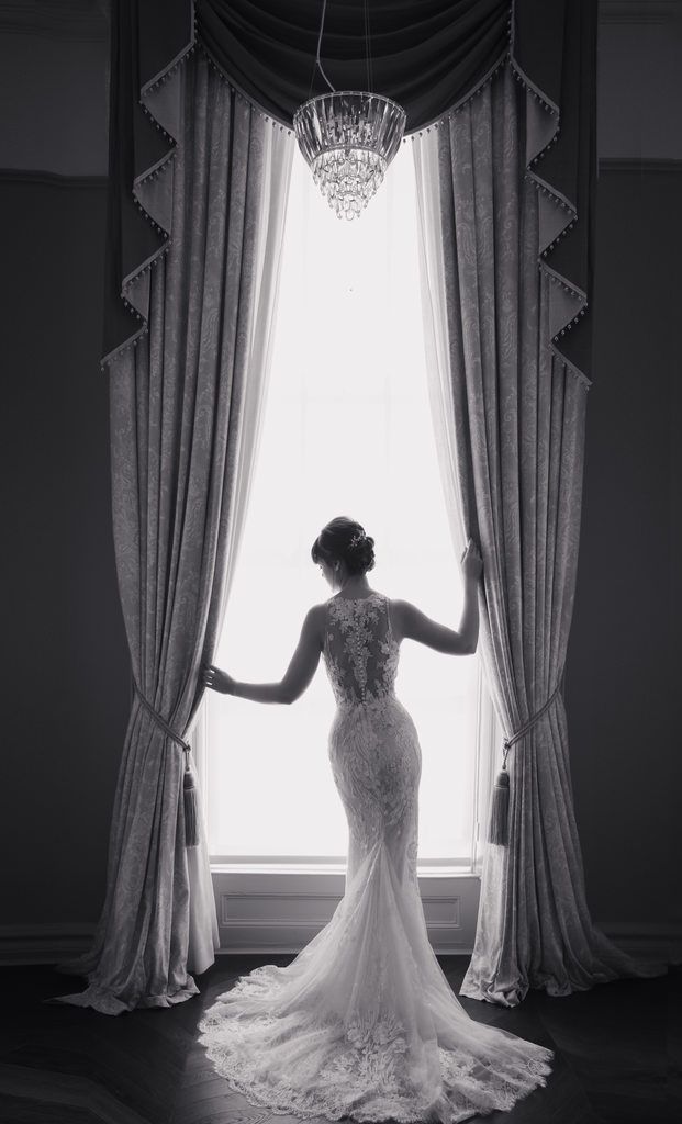 Silhouette of a bride in a lace gown standing by a grand window with elegant drapes and chandelier.
