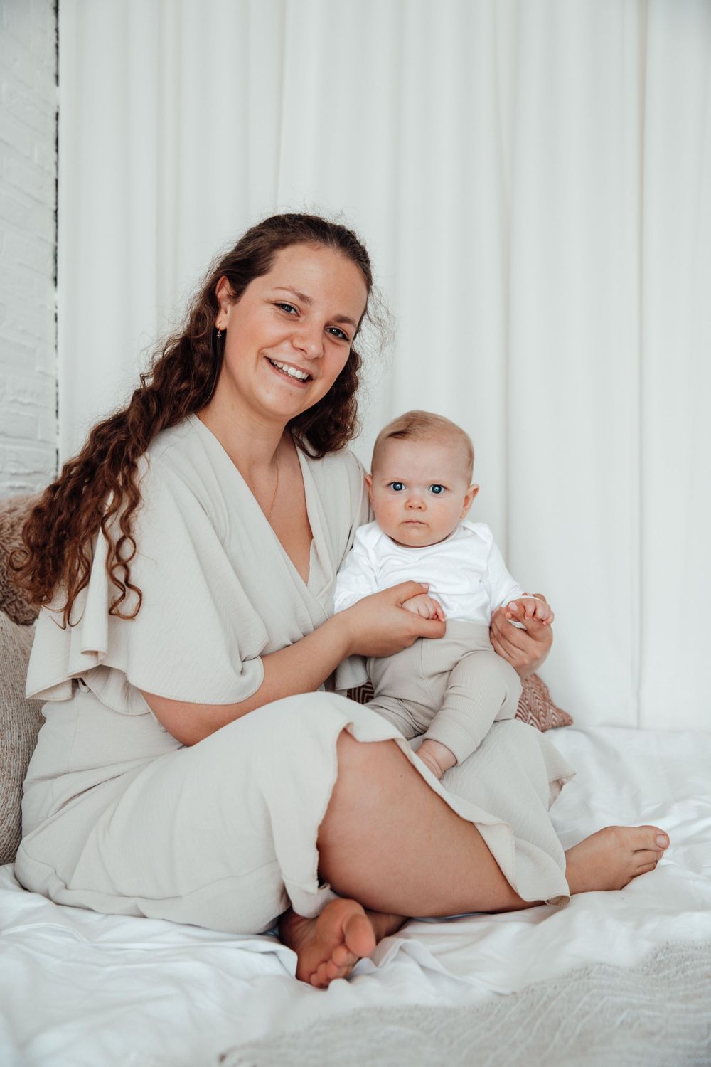 A mother and baby in matching cream colored outfits sit together on white bedding.