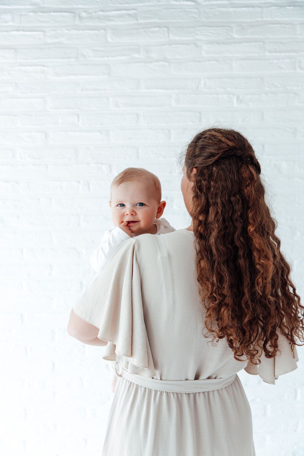 A baby peeks over someone's shoulder while being held, against a white brick wall background.
