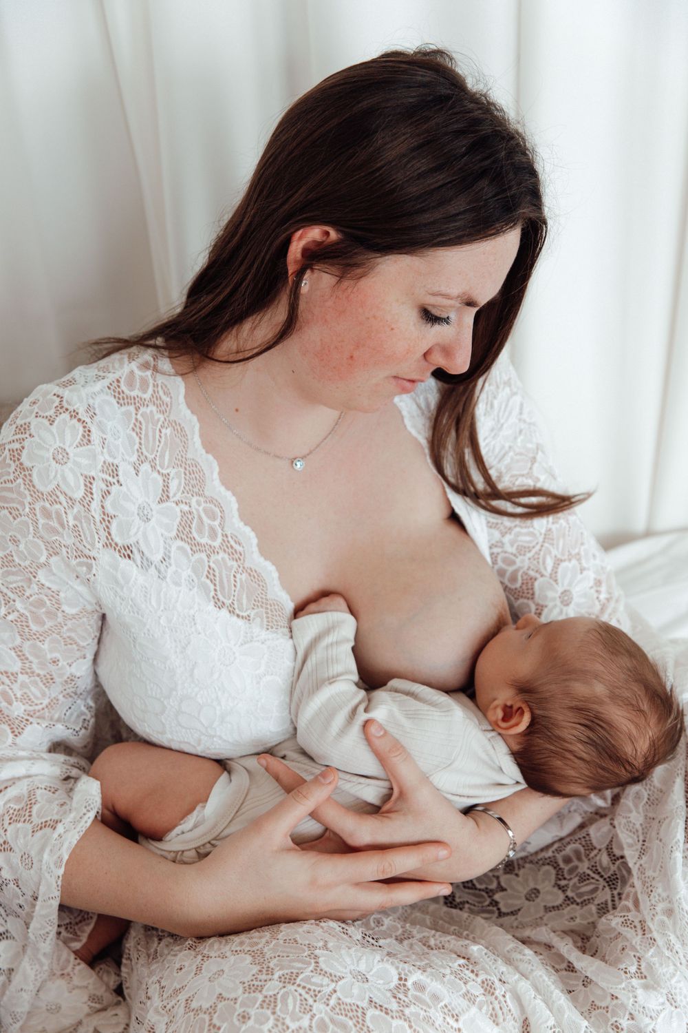 A peaceful breastfeeding moment captured in a white lace dress against light curtains.