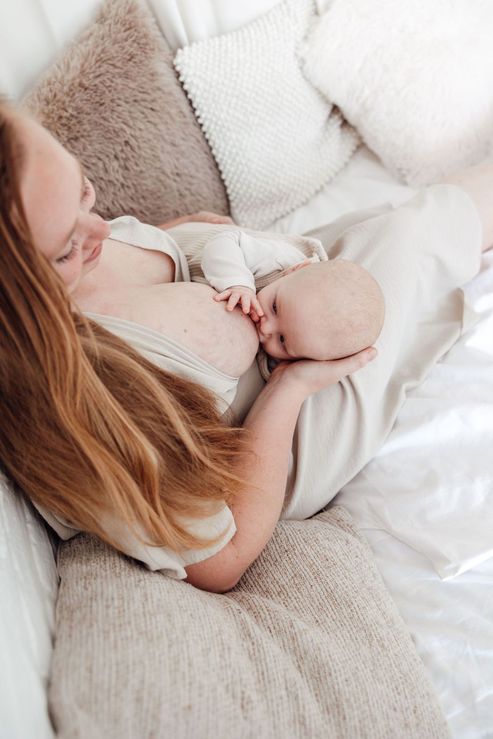 Intimate breastfeeding moment captured in soft natural light on white bedding.