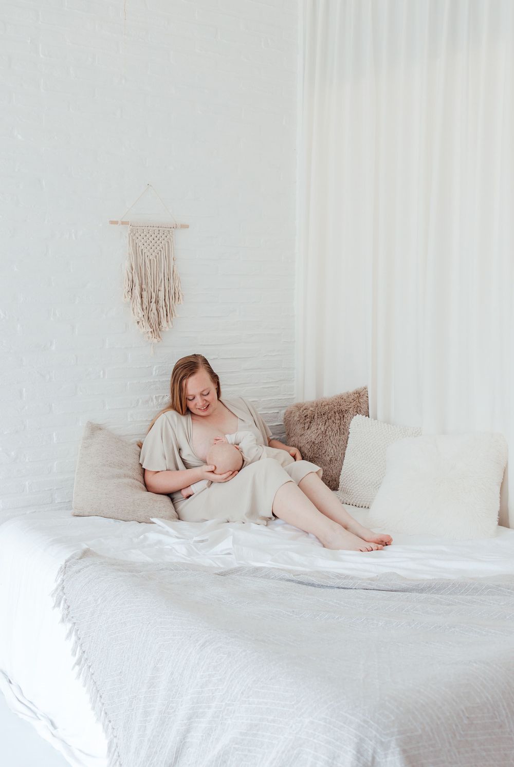Mother nursing baby in white bedroom with macrame wall decor.