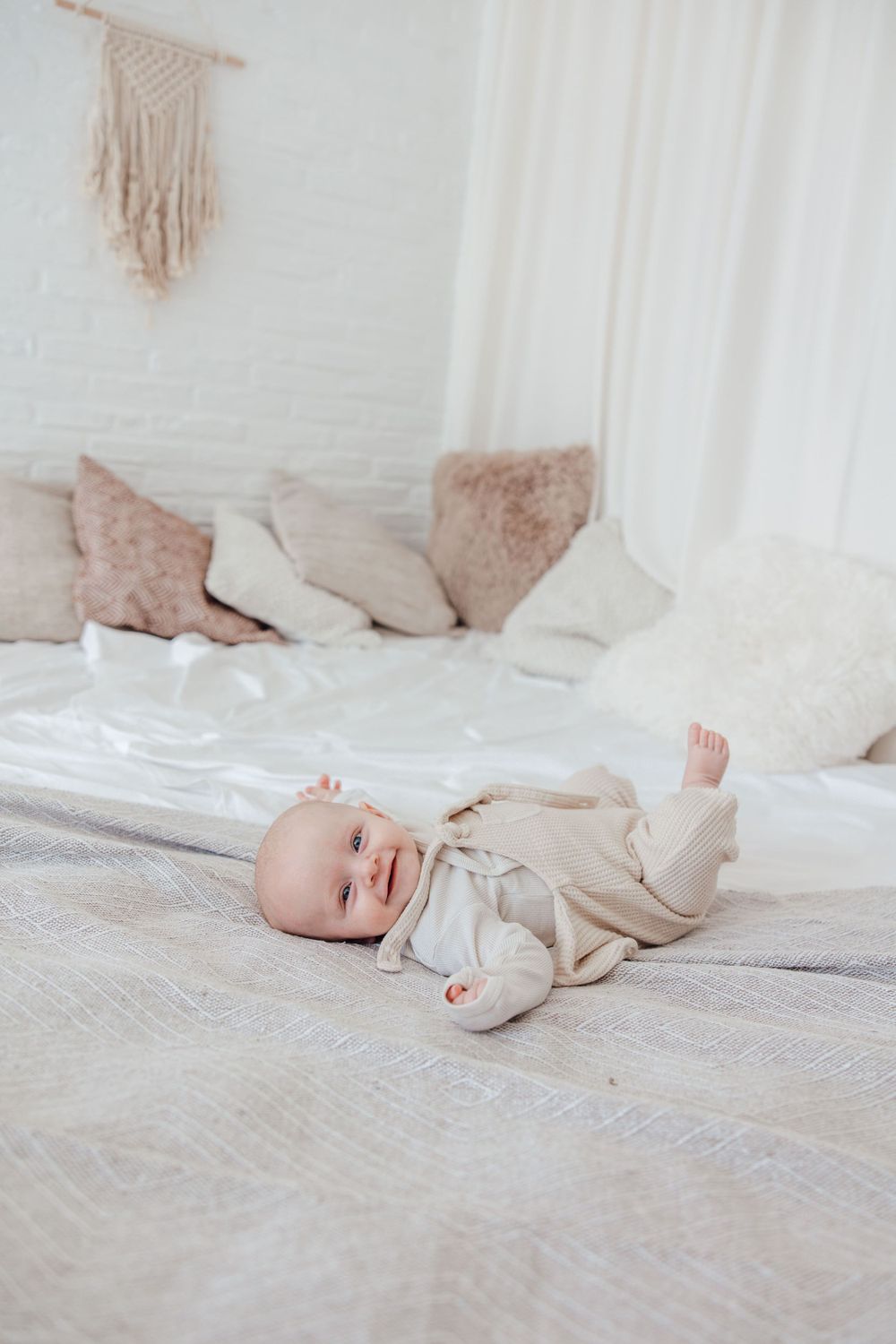 Baby in beige outfit lying on grey bedding with macrame wall decor.