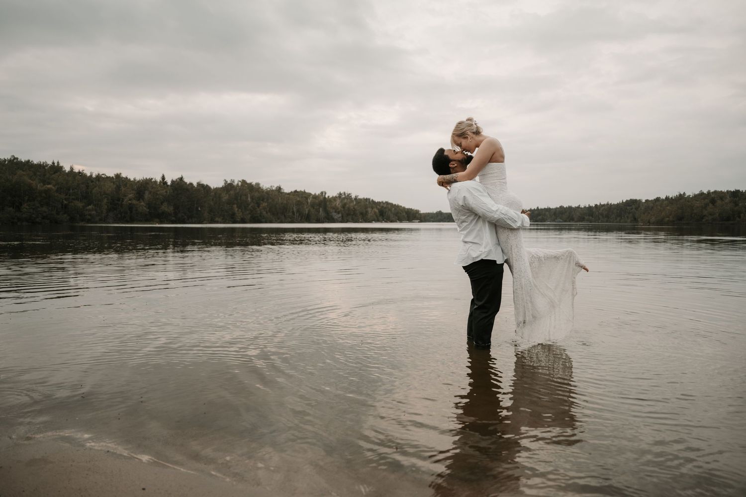 A couple embraces while standing in shallow lake water, with the bride's dress flowing in the breeze.