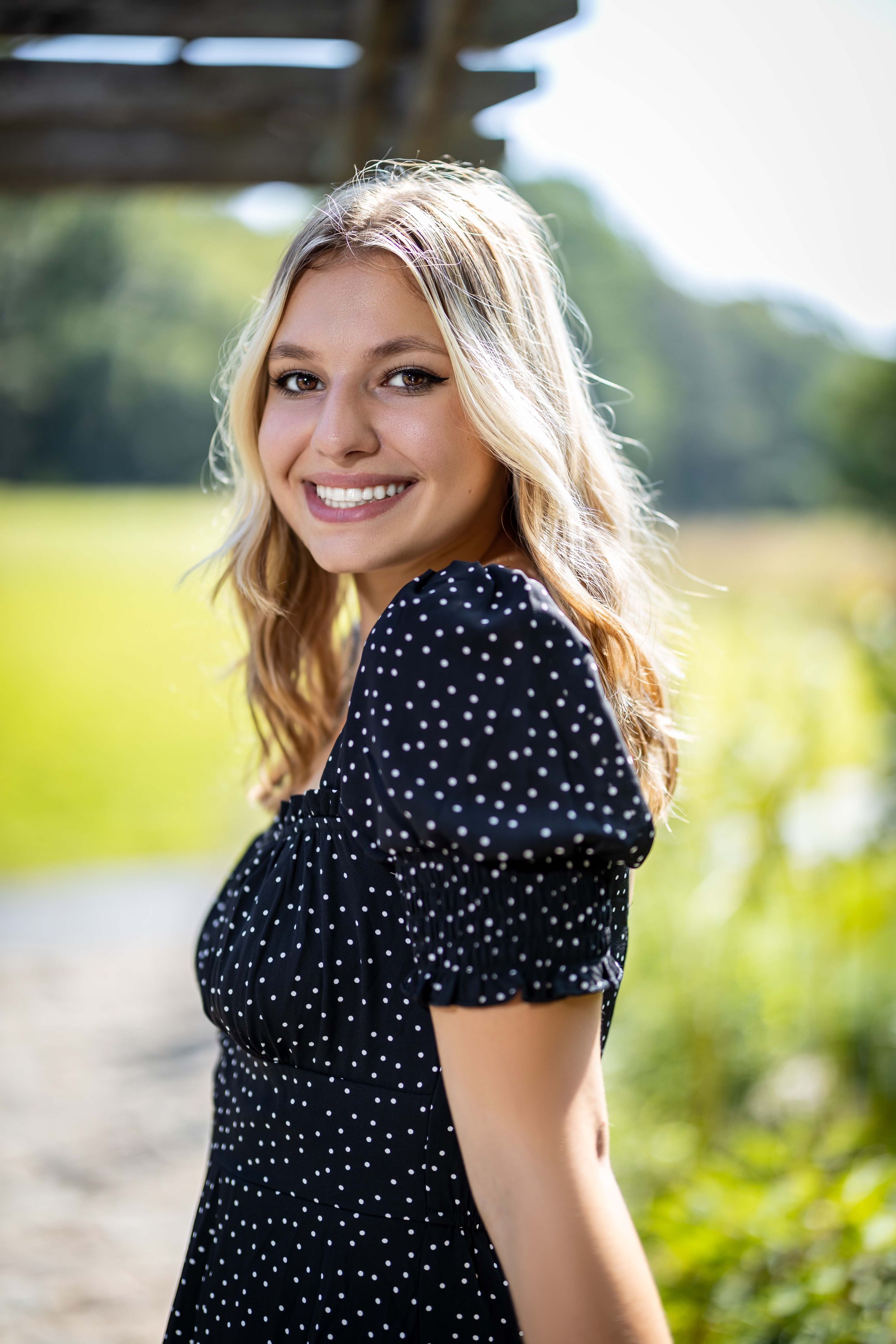 Person in polka dot dress smiles brightly in outdoor garden setting with natural lighting.