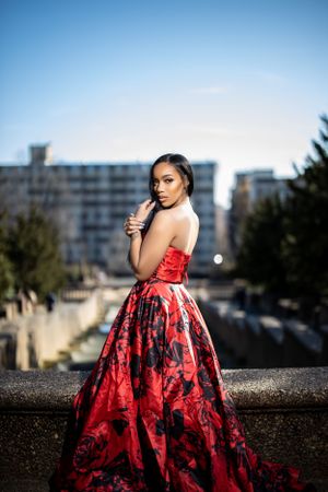 Person in dramatic red floral evening gown poses against urban backdrop at dusk.