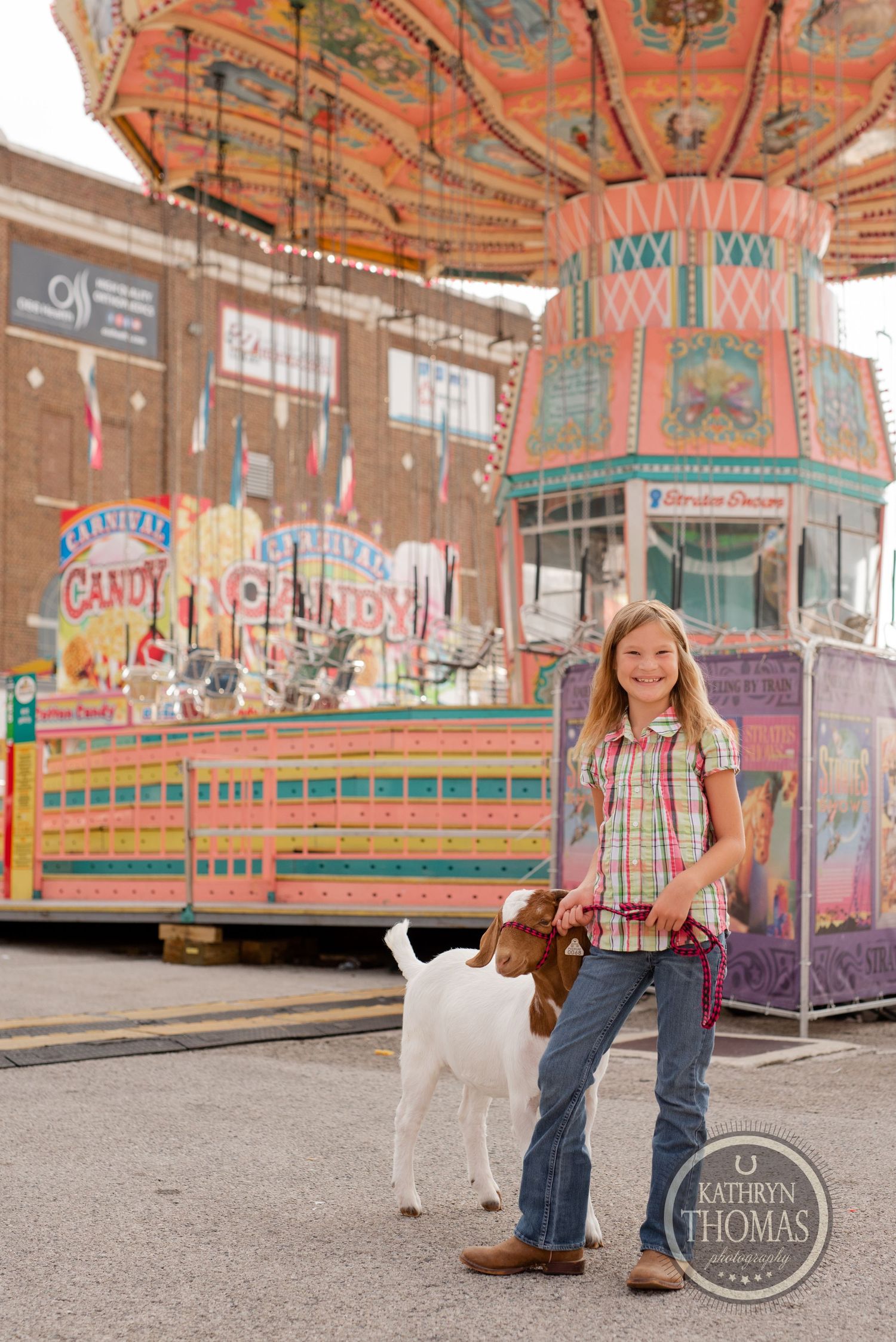 A young exhibitor leads a white goat past colorful fair concession stands.