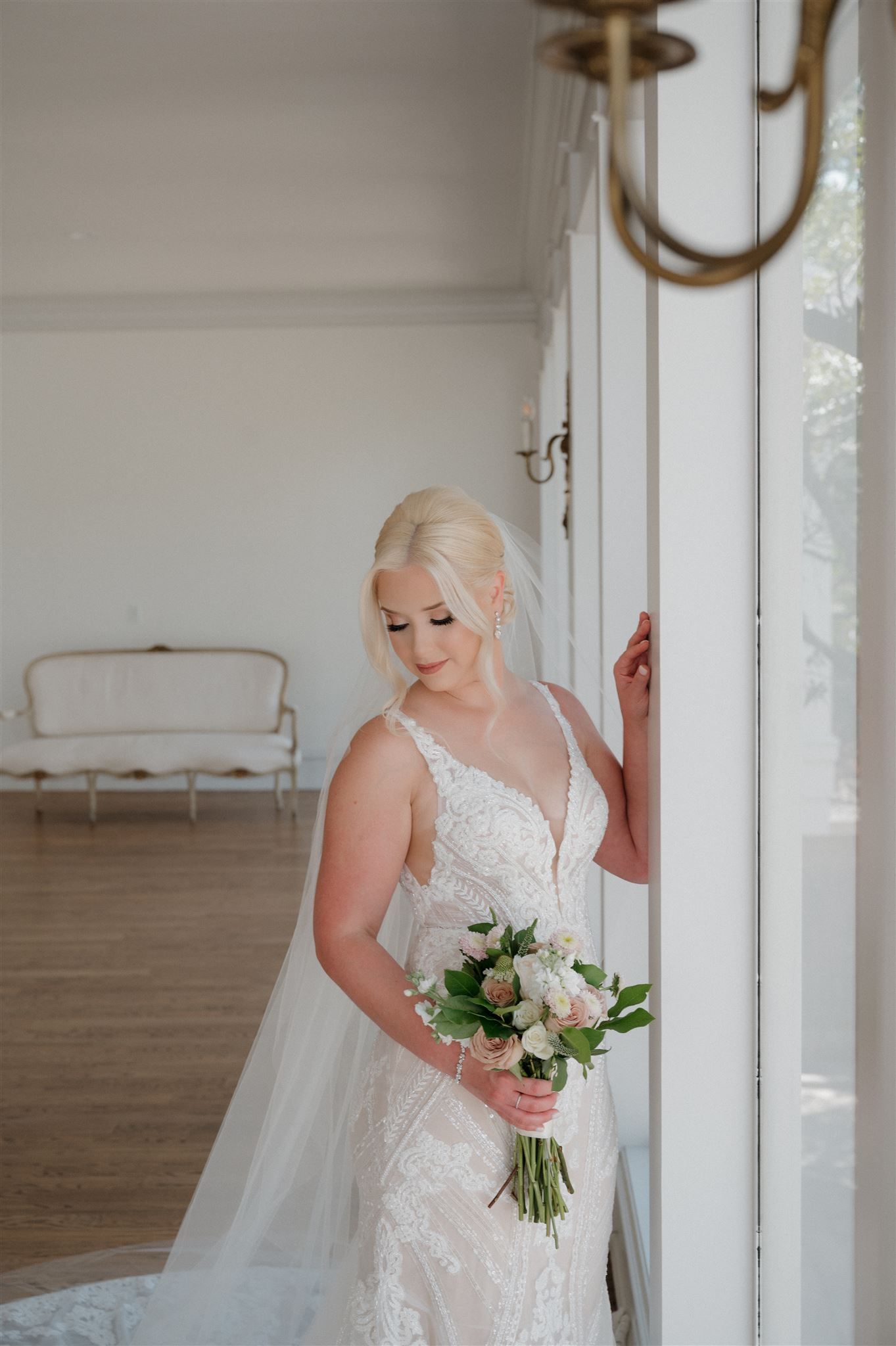 A bride in a fitted lace gown poses by a window holding a classic white and pink bouquet.