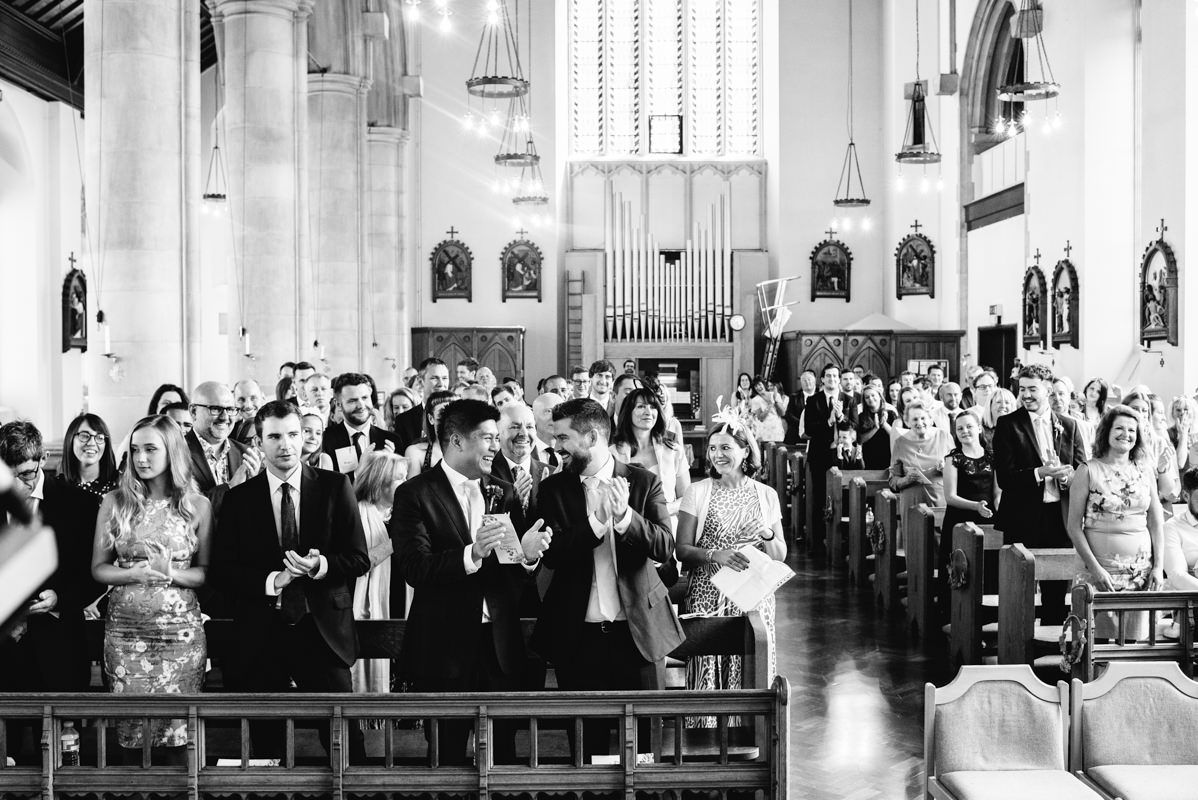 Wide angle black and white shot of wedding guests standing in church pews.