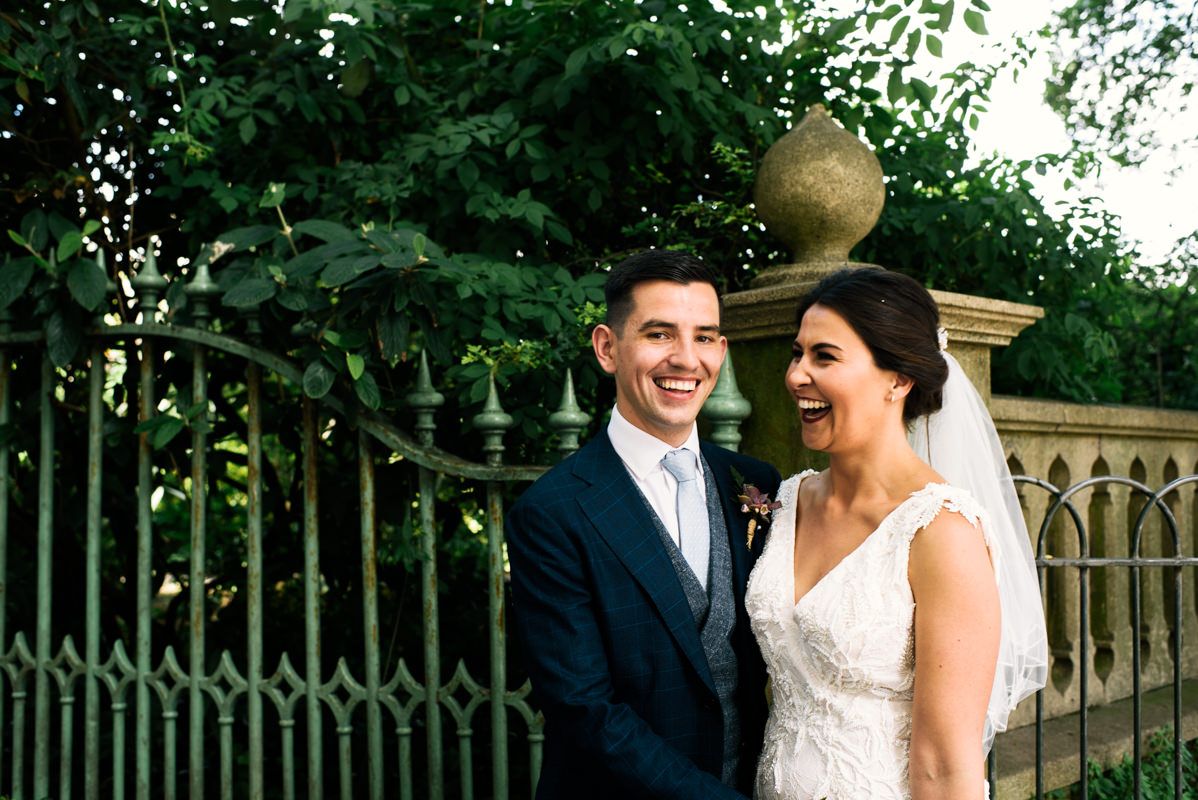 Joyful wedding portrait in garden setting with ornate architectural backdrop.