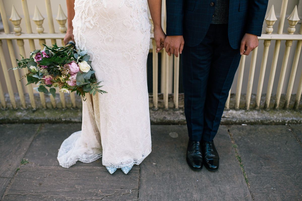 A wedding couple's feet showing a white lace dress and blue shoes next to formal black shoes.