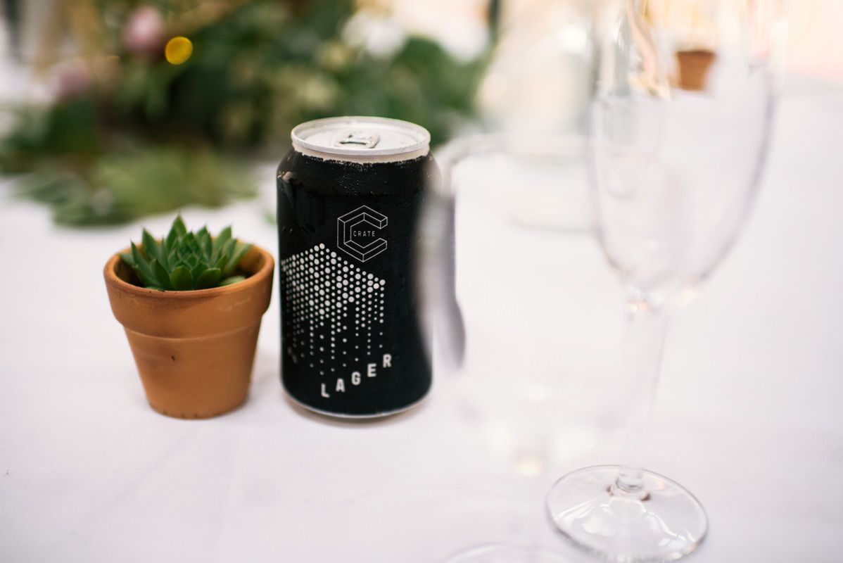 Black beverage can next to small potted succulent and champagne glass on white tablecloth.