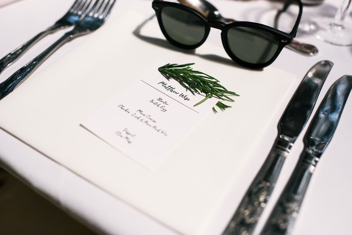 Place setting with menu card, sunglasses, and silverware on white tablecloth.