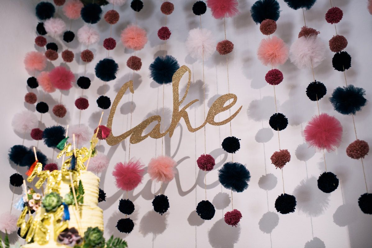 Decorative wall with black and pink pom poms and gold 'Cake' text for wedding reception.