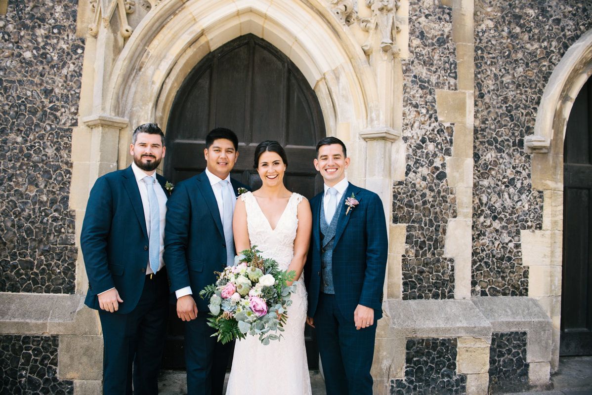 Wedding portrait of couple with groomsmen in navy suits against stone church facade.