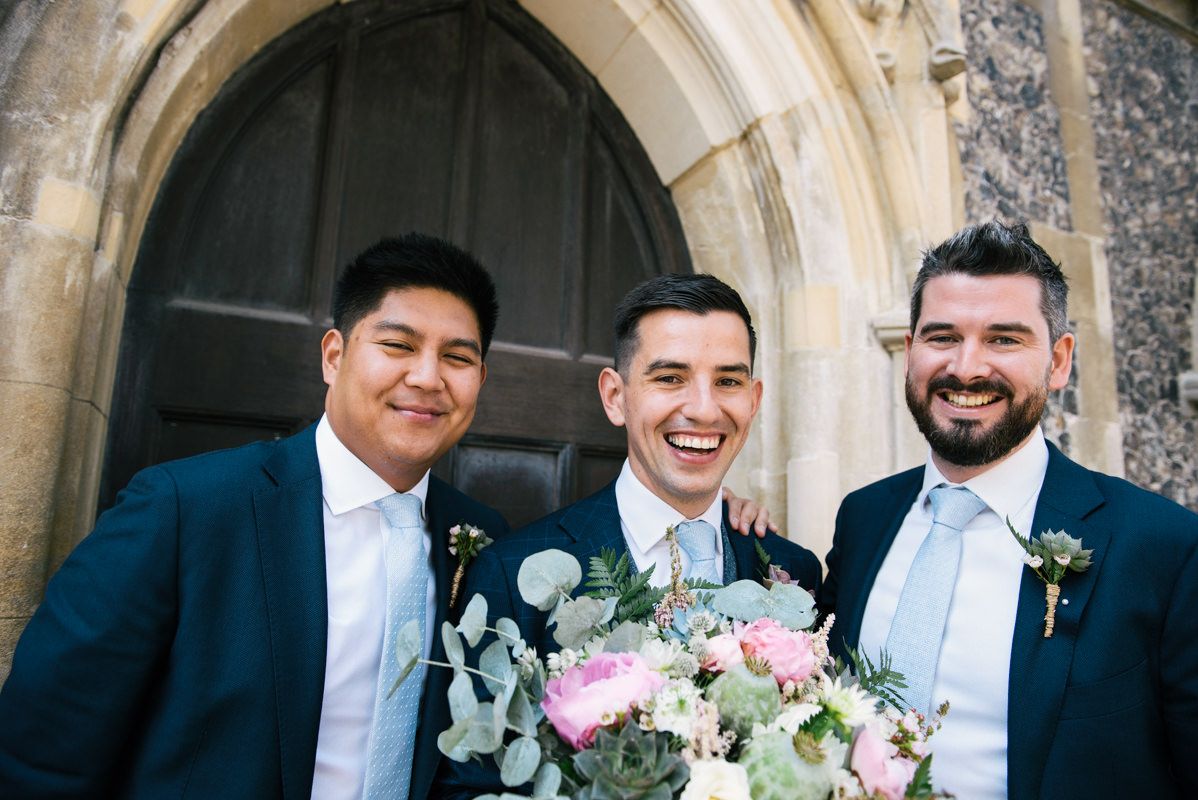 Three groomsmen in navy suits pose outside a historic stone church entrance.