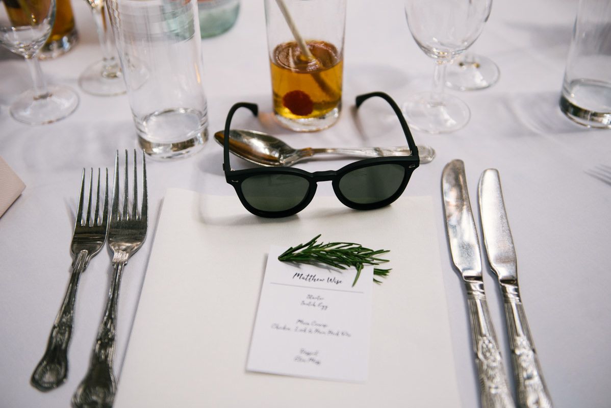 Wedding place setting with sunglasses, menu card, and elegant silverware on white tablecloth.
