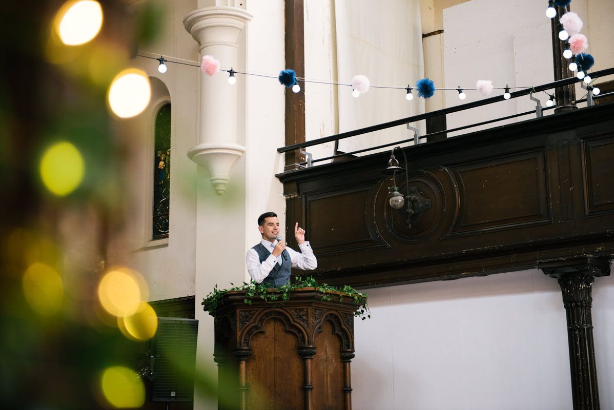 Two people give speeches from an ornate wooden balcony during a wedding reception.