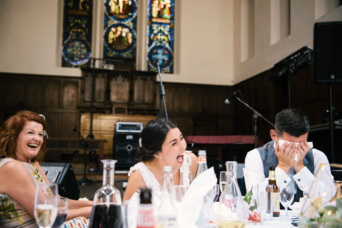 Wedding party members react with laughter during reception speeches in a church venue.