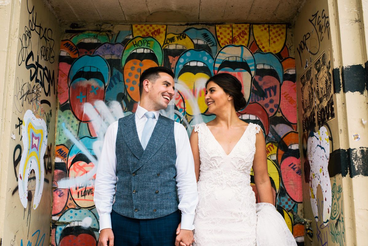 Wedding couple shares a joyful moment against vibrant graffiti wall covered in colorful face designs.