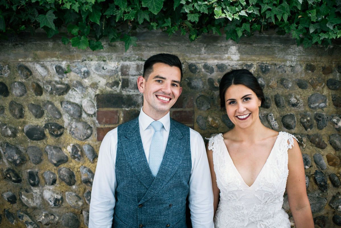 Newlyweds stand against stone wall with greenery for outdoor wedding portrait.