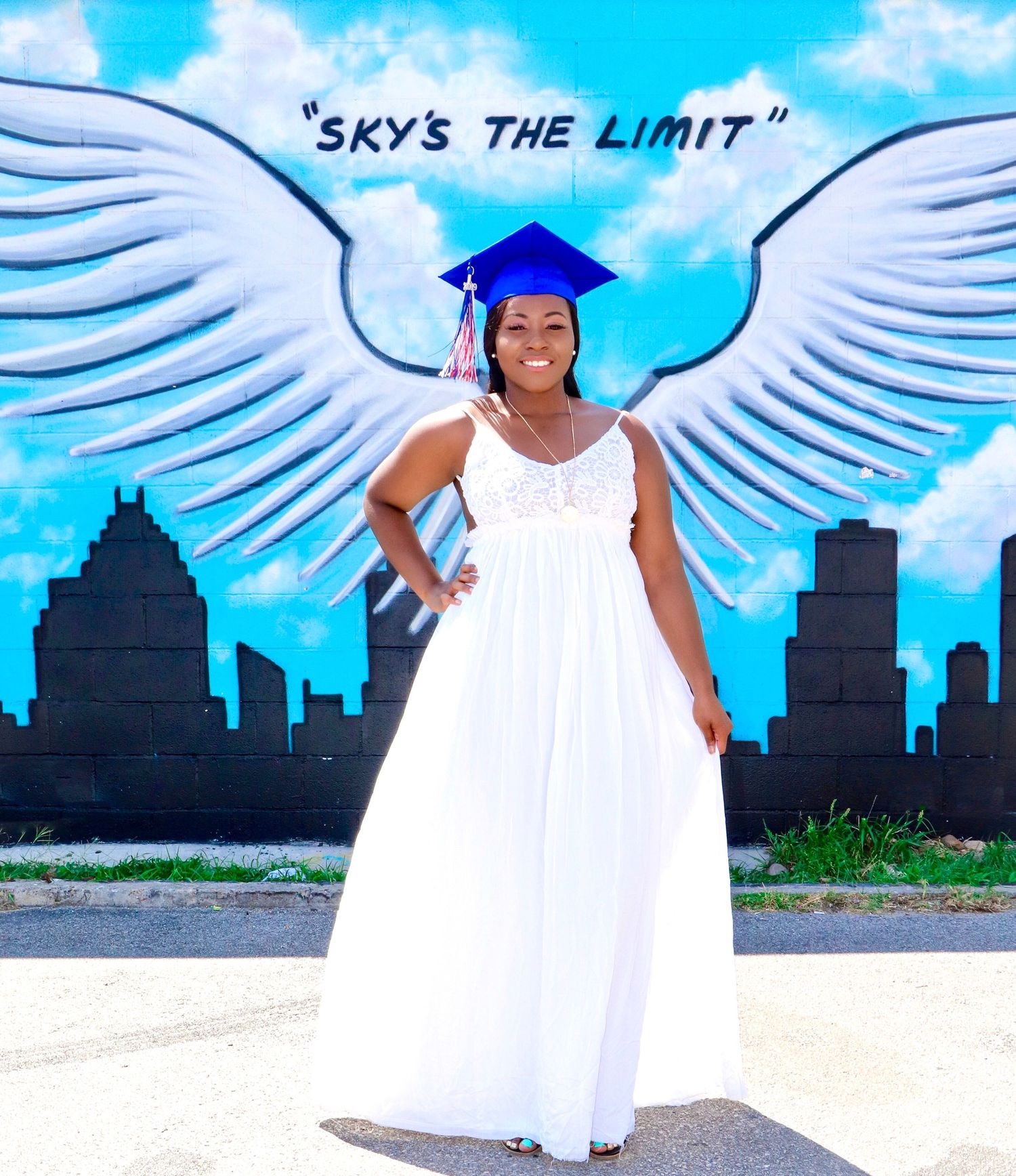 Graduate in white dress poses in front of angel wings street art with city skyline.