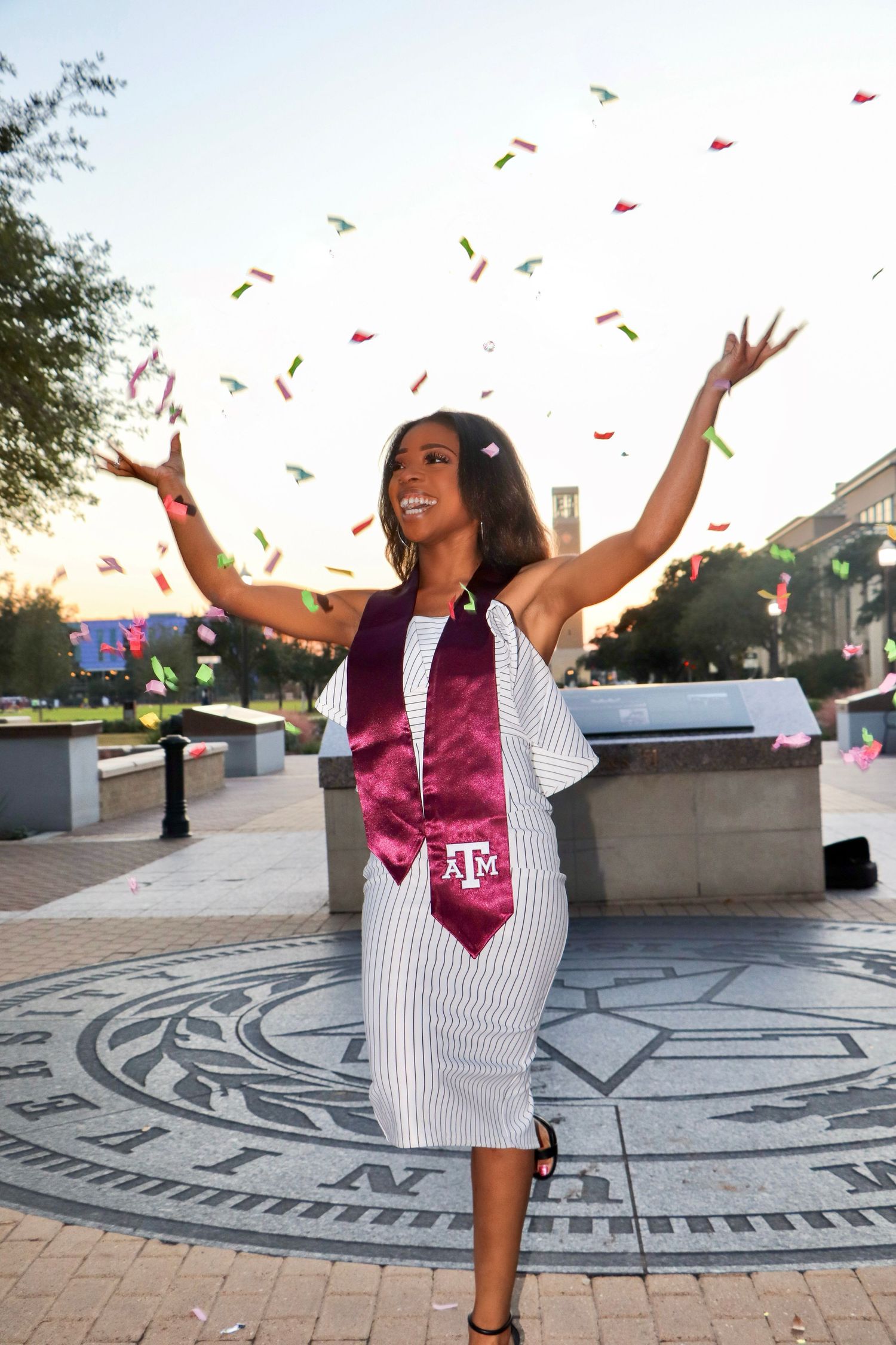 Graduate in white dress and purple stole celebrating with confetti on university campus.