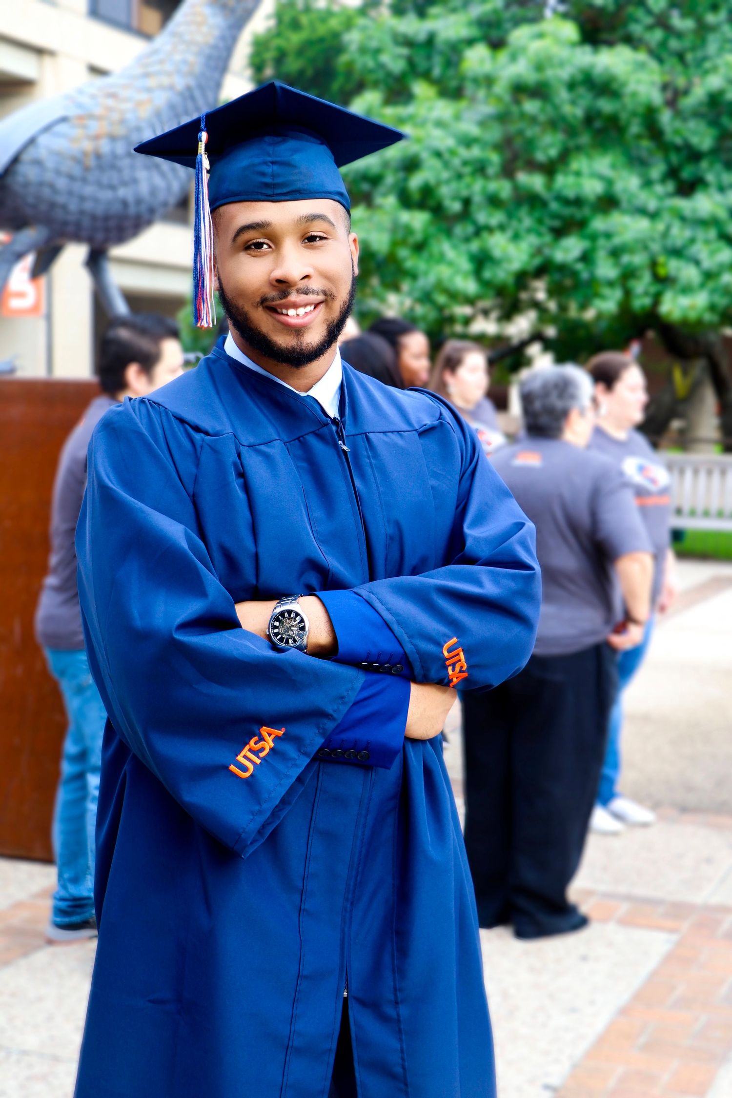 Graduate in traditional blue cap and gown smiling outdoors on campus.
