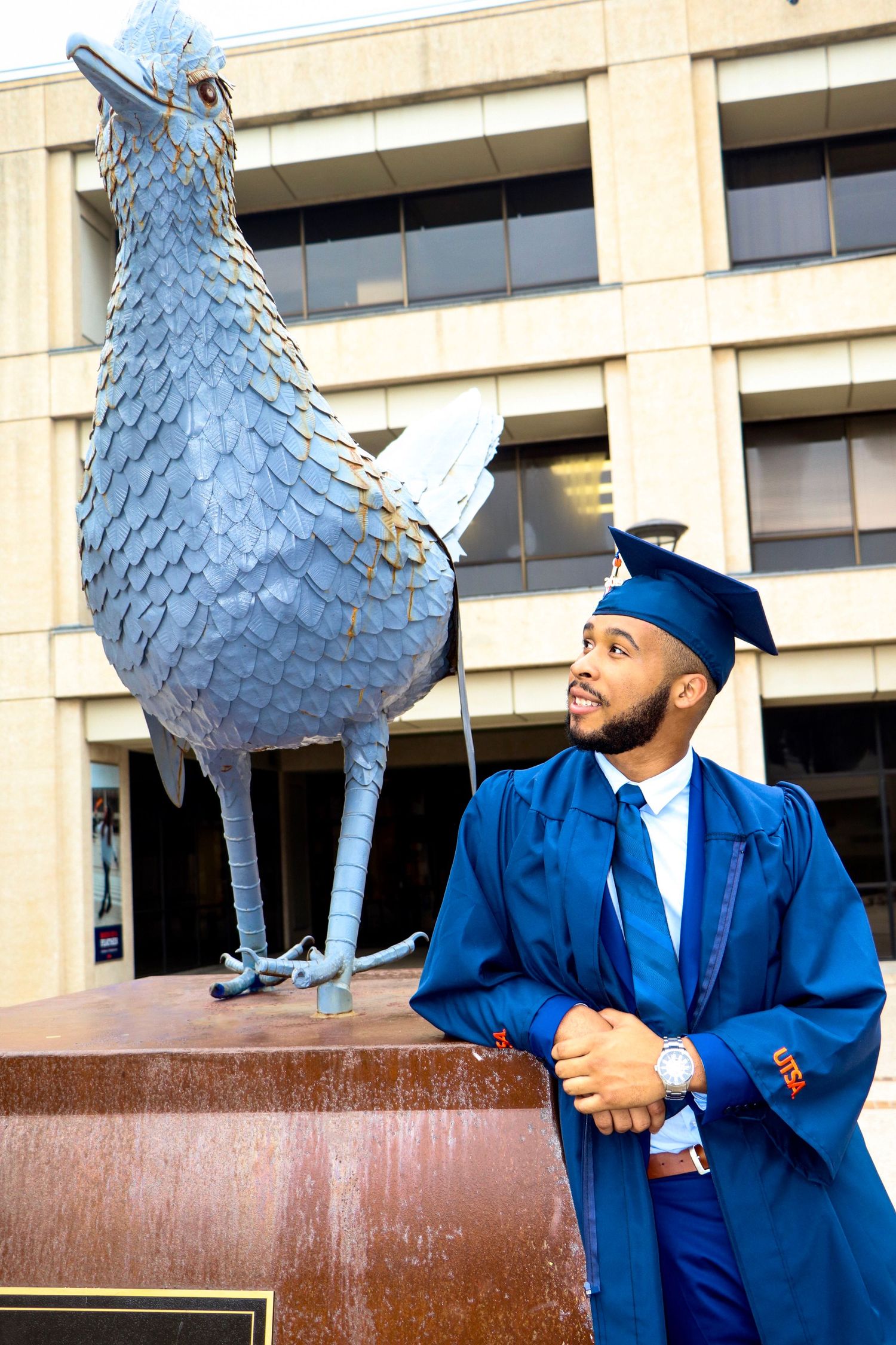 Graduate in blue academic regalia poses next to large metal bird sculpture.