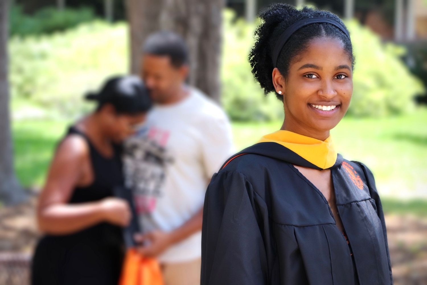 A graduate in academic regalia with a yellow stole poses outdoors on a sunny day.