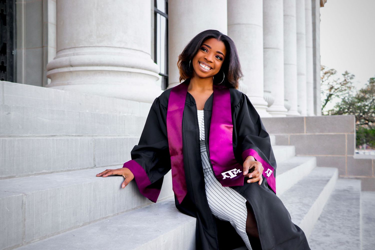 Graduate in black and purple graduation regalia smiling on white column steps.