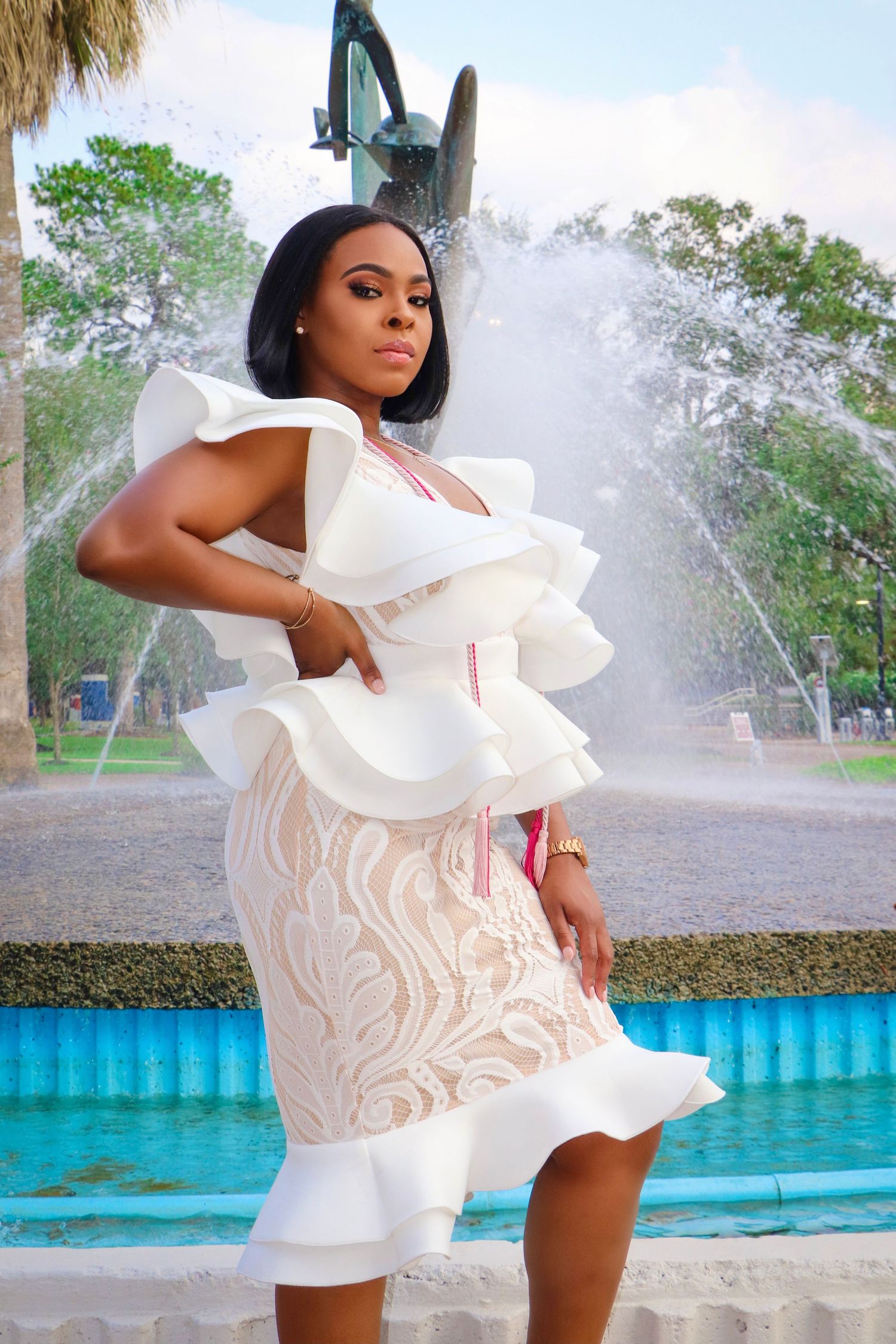 Fashion photo near fountain showing white textured dress with ruffled details and architectural background.