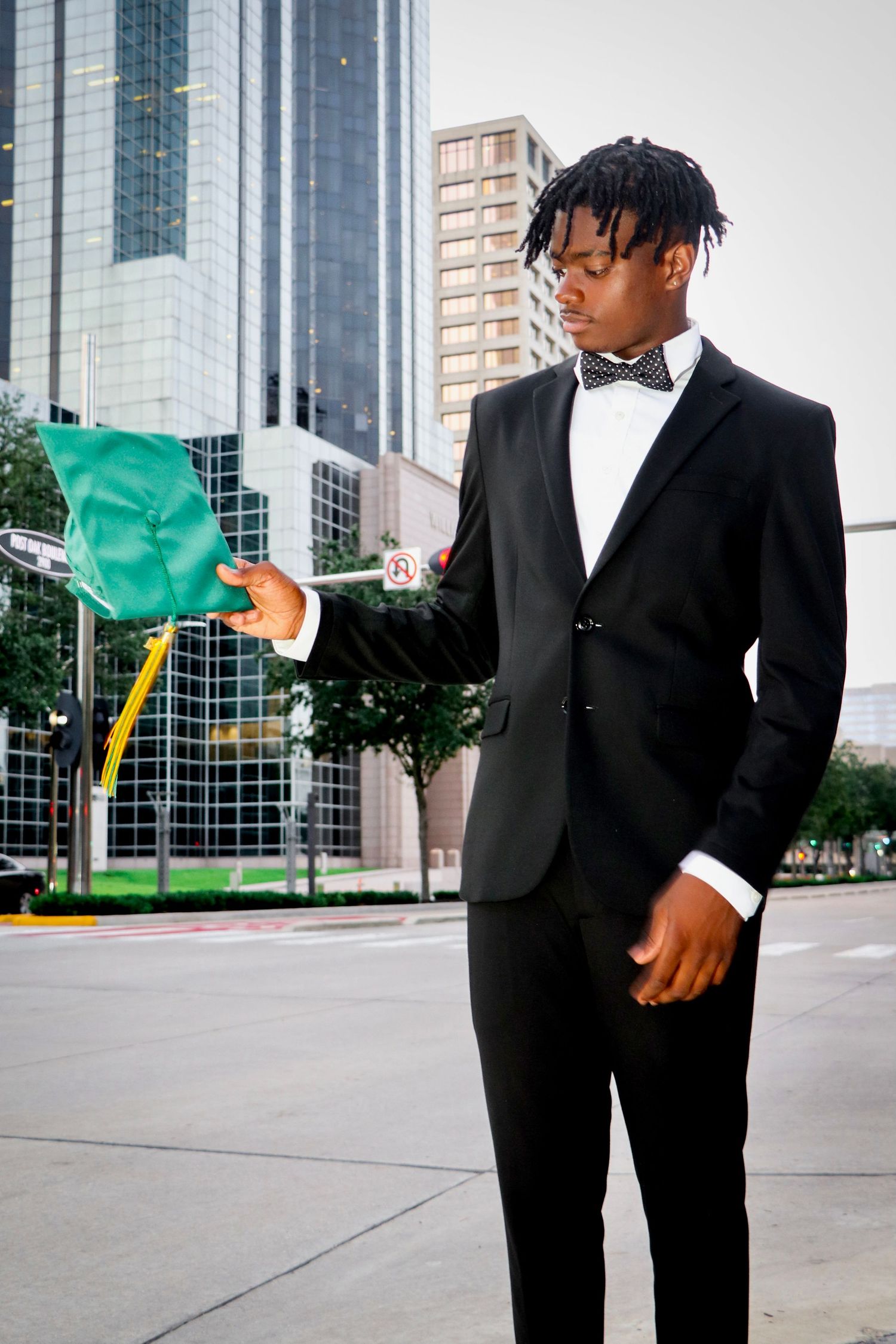 Graduate in formal black tuxedo and bowtie holding green flag in urban setting.