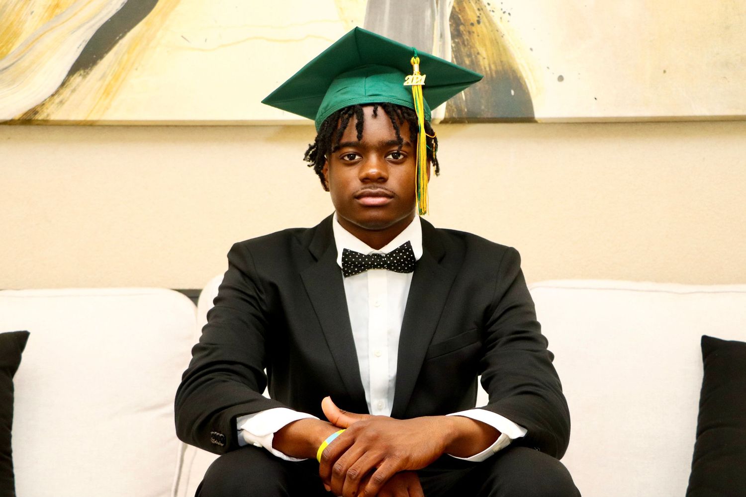 Graduate wearing green cap and black tuxedo sitting formally on white couch.