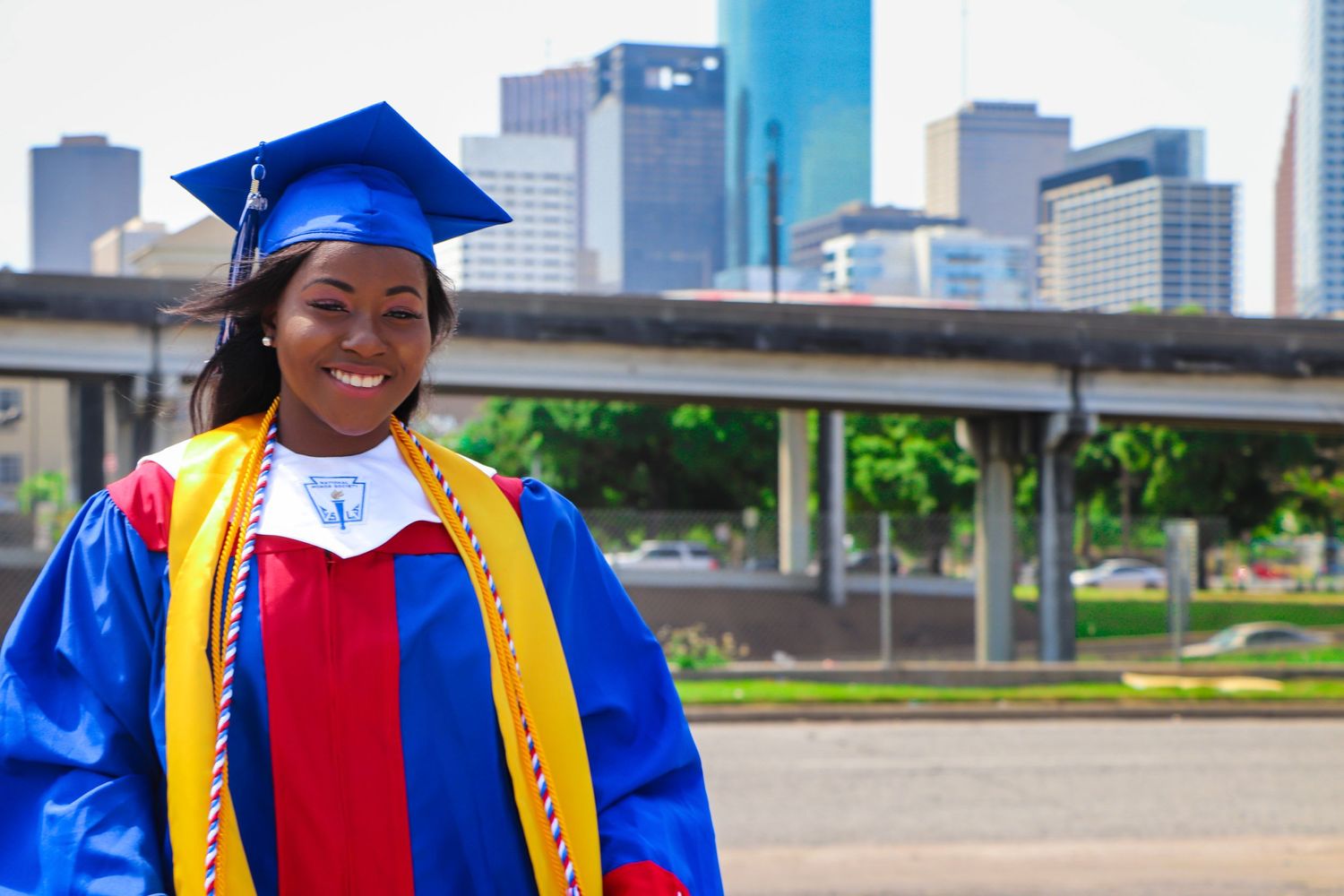 Graduate in blue and yellow regalia poses with city skyline in background.