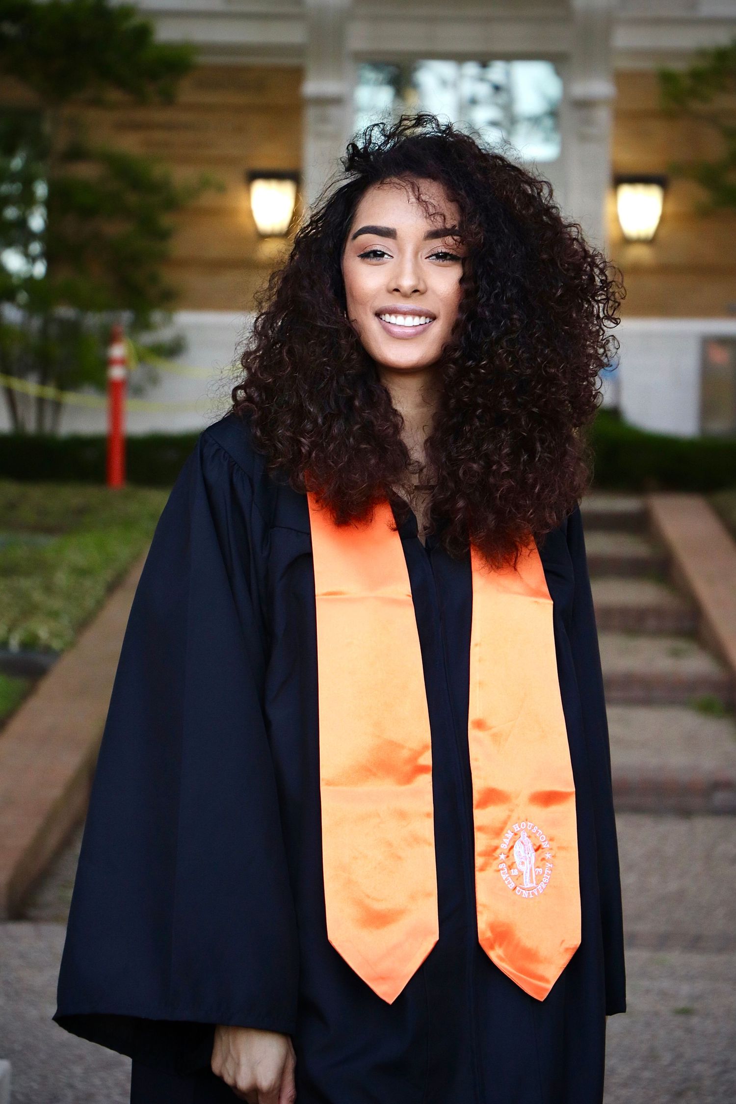Graduate in navy cap and gown with orange stole standing outdoors.