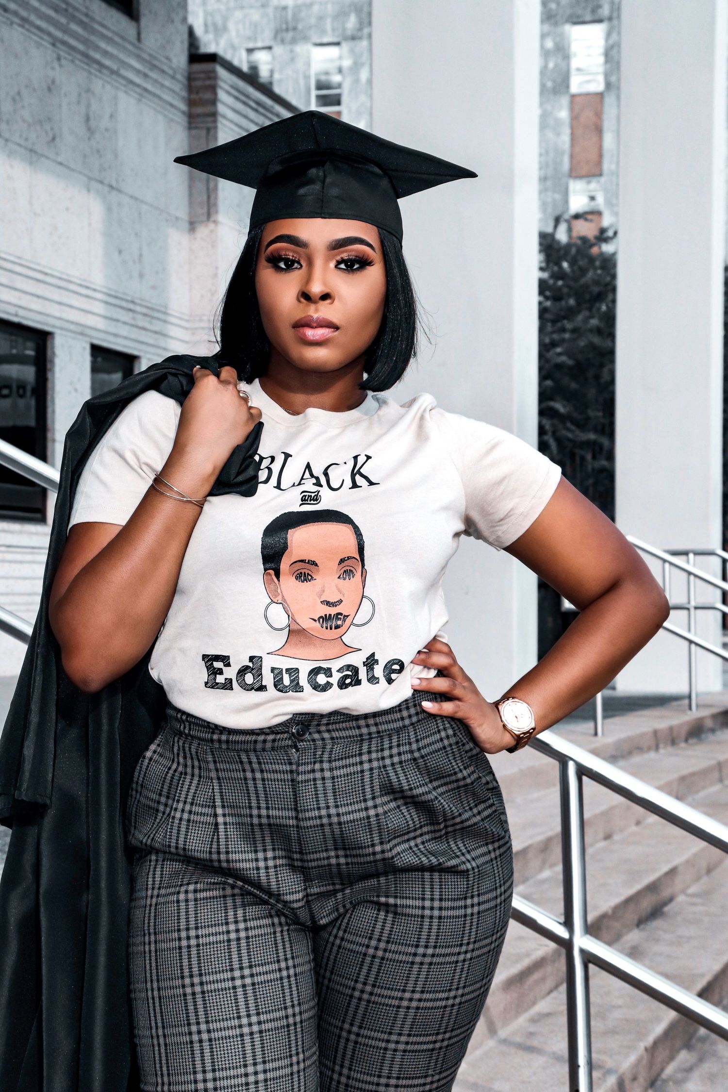 Graduate in cap and gown wearing Black Educate t-shirt on campus steps.