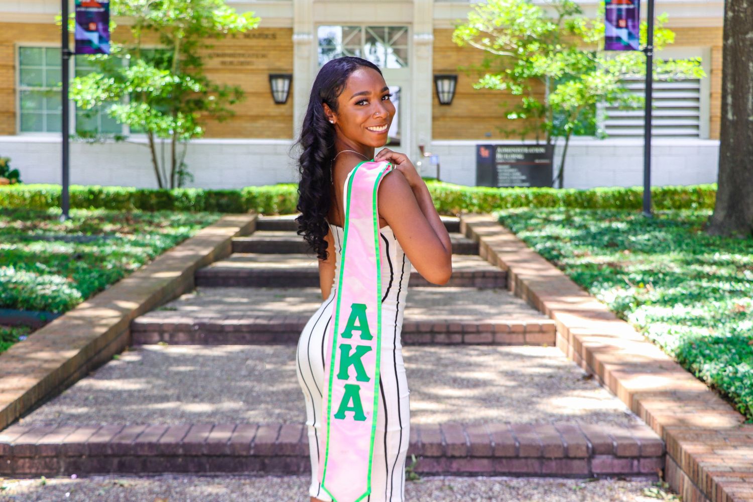 Student in white and green striped dress poses on campus steps surrounded by landscaping.
