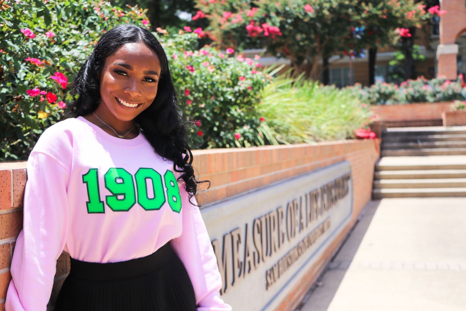 Student in pink sweatshirt with green 1908 numbers smiles near campus signage.