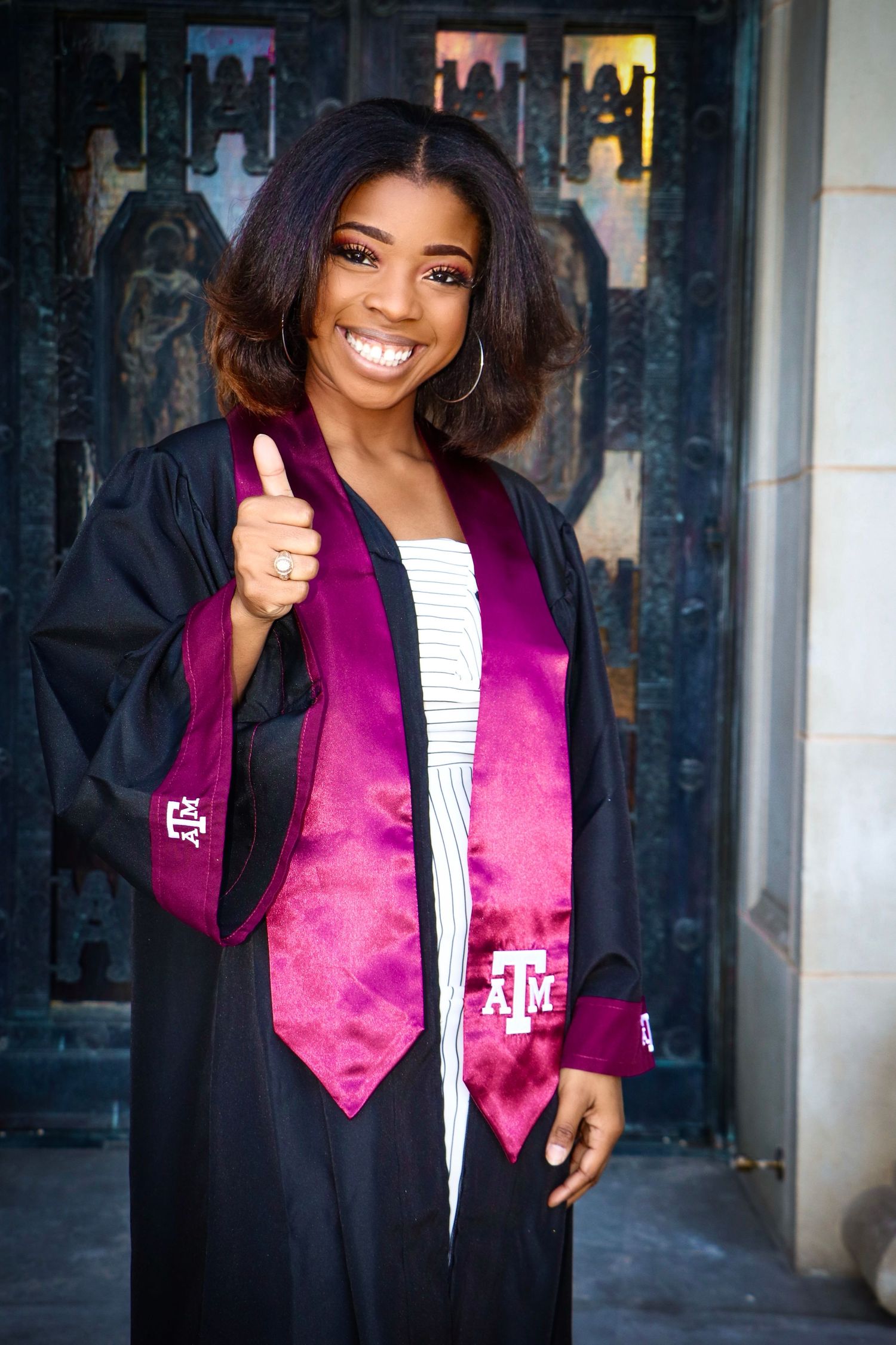 Graduate in black gown with purple stole giving thumbs up in front of decorative doorway.