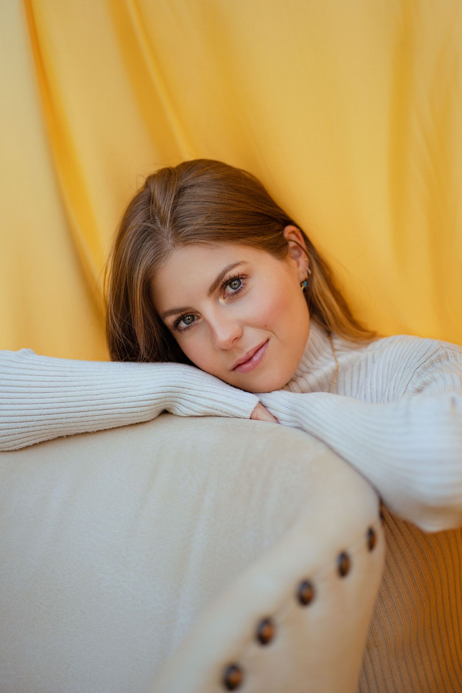 Person in white sweater resting on beige couch against yellow backdrop.