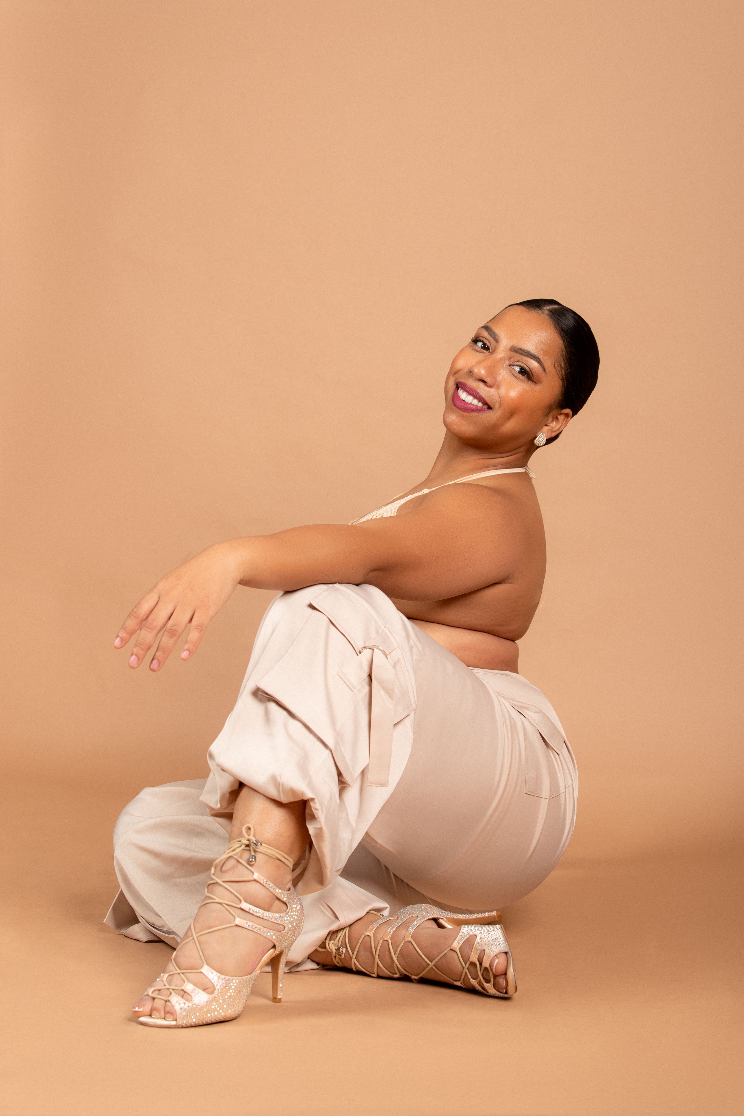 A fashion portrait of a model in cream-colored outfit and strappy sandals against a peach background.