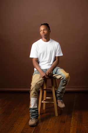 Portrait of person in casual attire sitting on wooden stool against brown backdrop.